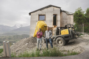 Deux personnes se tiennent devant une maison en construction abandonnée, avec des baies vitrées inachevées et une pelle mécanique jaune à l'avant. La maison est située dans une région montagneuse avec une vue dégagée sur la vallée du Rhône, Valais, Suisse.