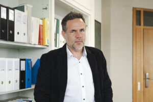 Un homme en chemise blanche et blazer noir se tient devant une bibliothèque blanche remplie de classeurs fédéraux à l'intérieur d'une pièce.