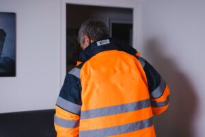 Un homme vu de dos portant une veste de travail orange et grise dans une pièce à l'intérieur d'une maison.